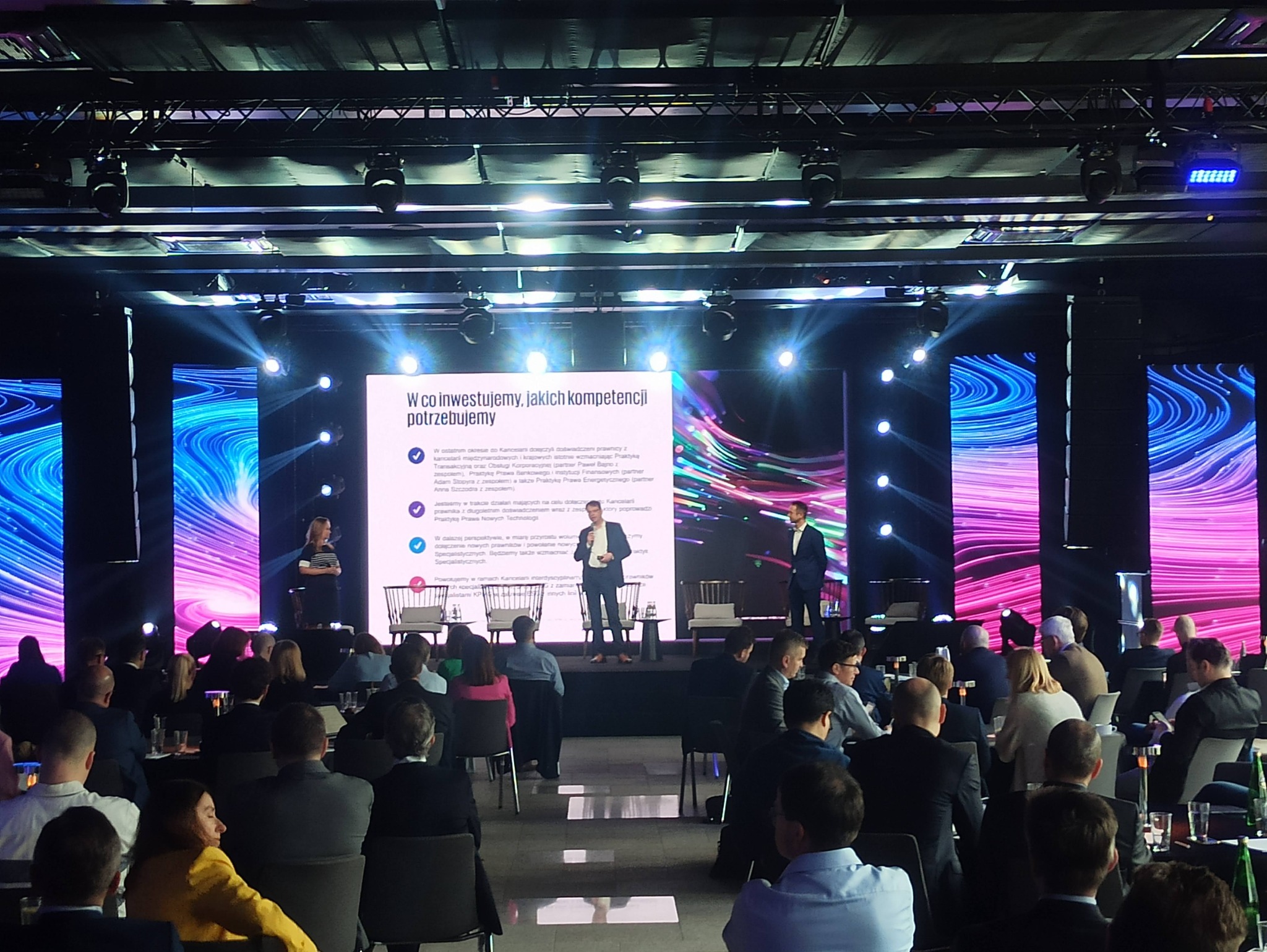 Konferencja z prelegentami na scenie oświetlonej reflektorami i z ekranami LED wyświetlającymi dynamiczne grafiki, widownia siedząca w rzędach.