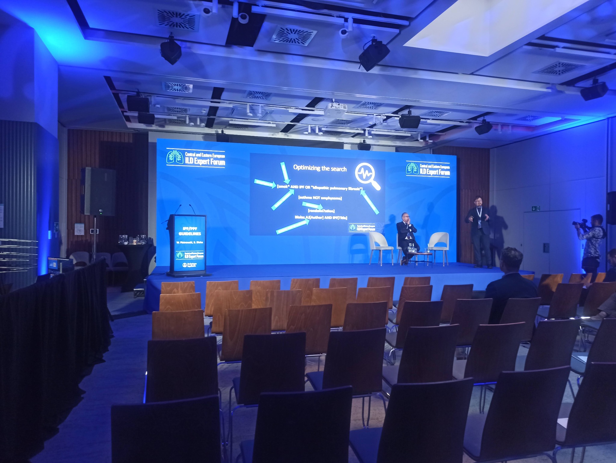 Sala konferencyjna z niebieskim oświetleniem, scena z ekranem wyświetlającym prezentację o tematyce medycznej, rząd krzeseł dla publiczności, prelegenci na scenie