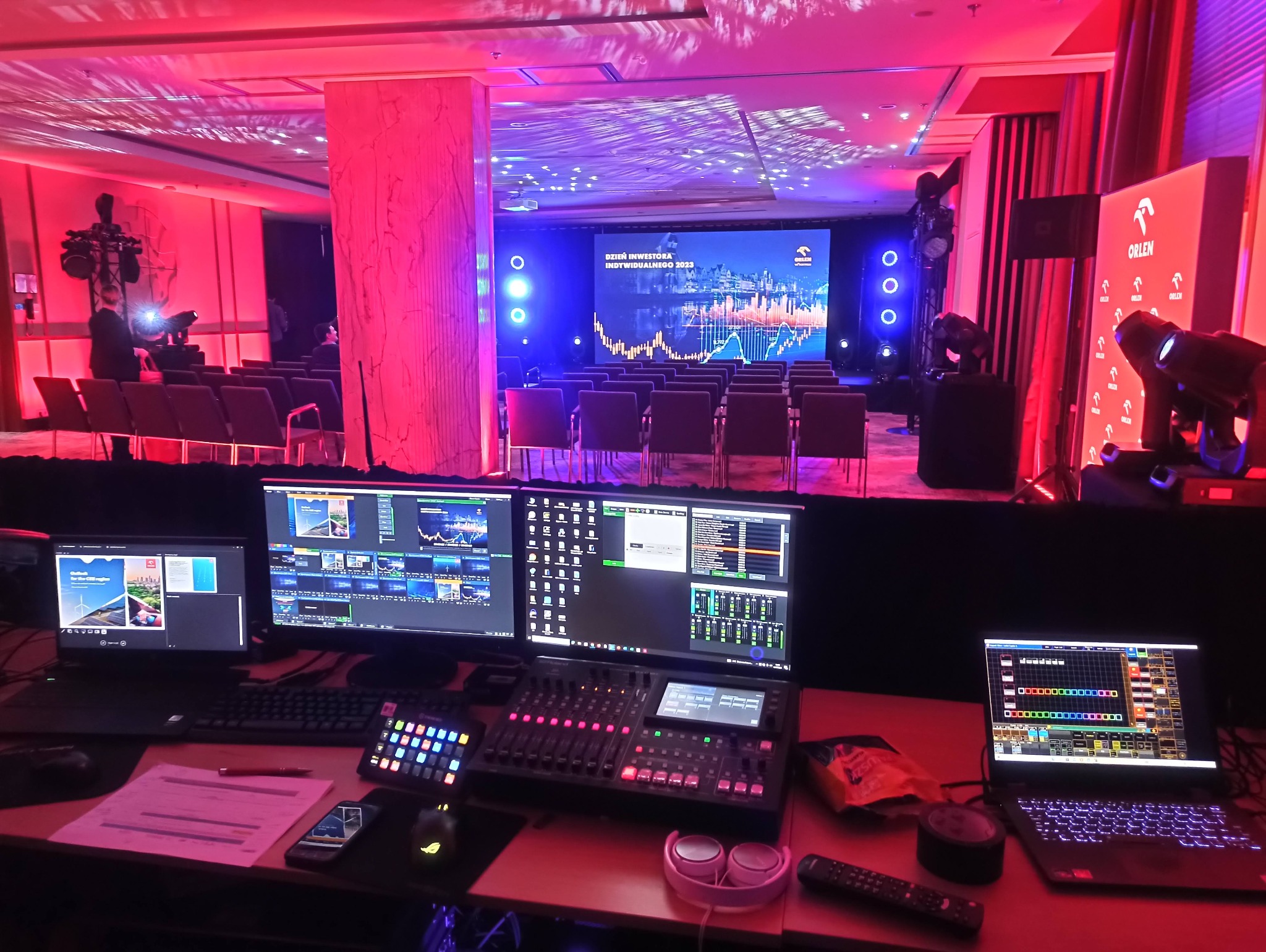 Stanowisko realizacji nagłośnienia i oświetlenia podczas konferencji, widoczne monitory, mikser audio, laptop z oprogramowaniem sterującym, reflektory sceniczne i ekran z prezentacją.