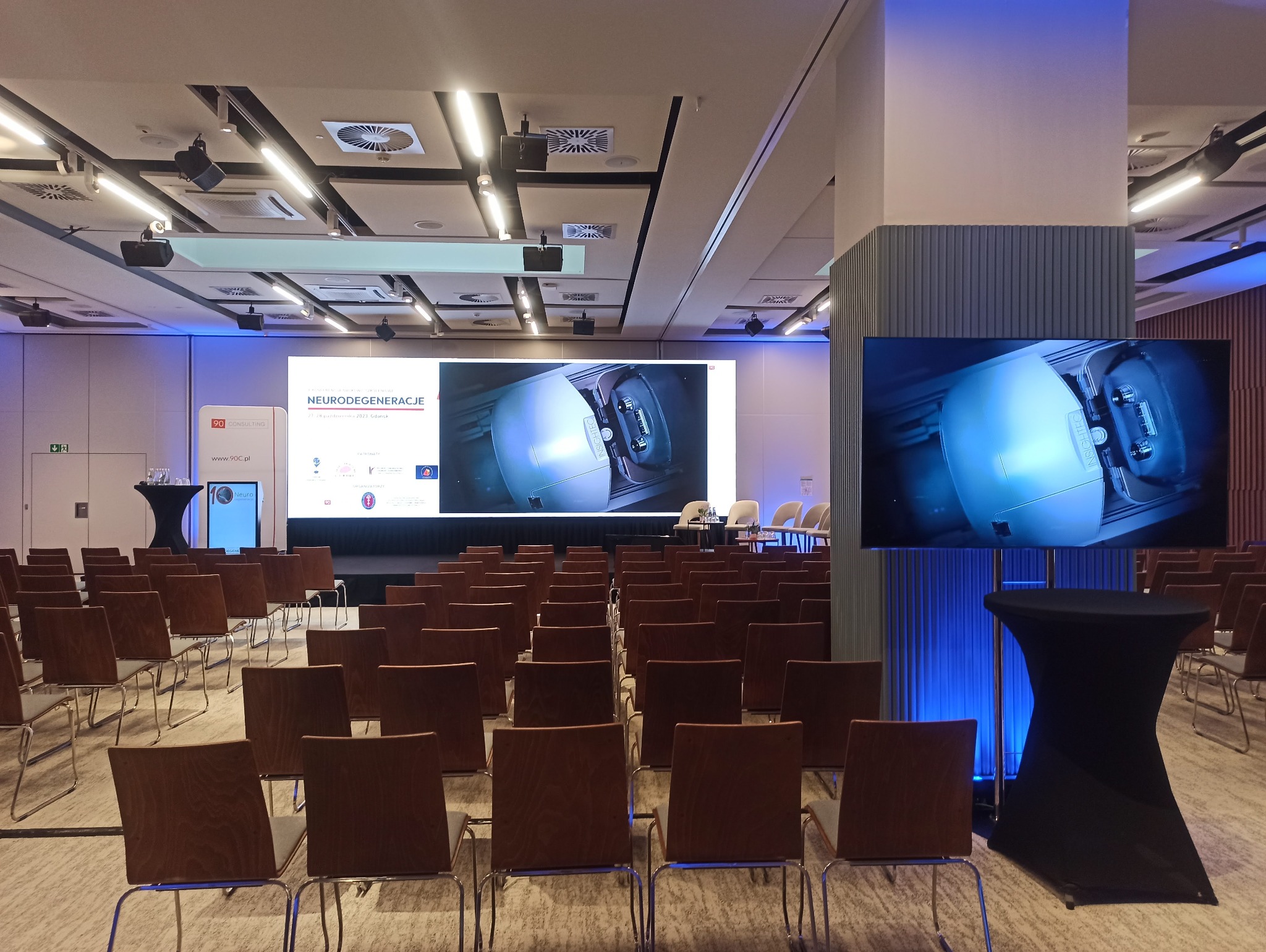 Sala konferencyjna z rzędami krzeseł ustawionych przed dużym ekranem wyświetlającym urządzenie medyczne i mniejszym ekranem po prawej stronie, oświetlona niebieskim światłem.