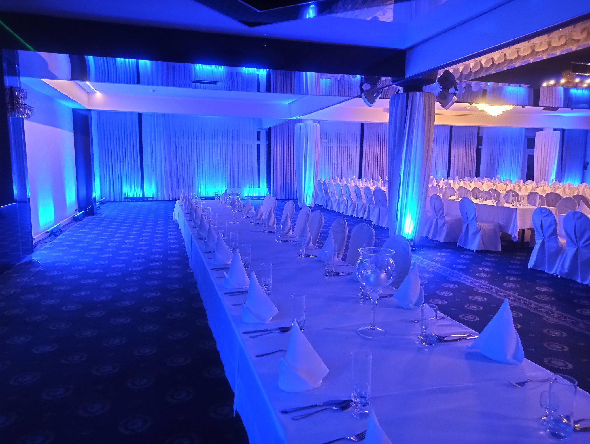 Sala bankietowa z długimi stołami nakrytymi białymi obrusami, białymi serwetkami ułożonymi w trójkąty, szklankami i sztućcami, oświetlona niebieskim światłem.