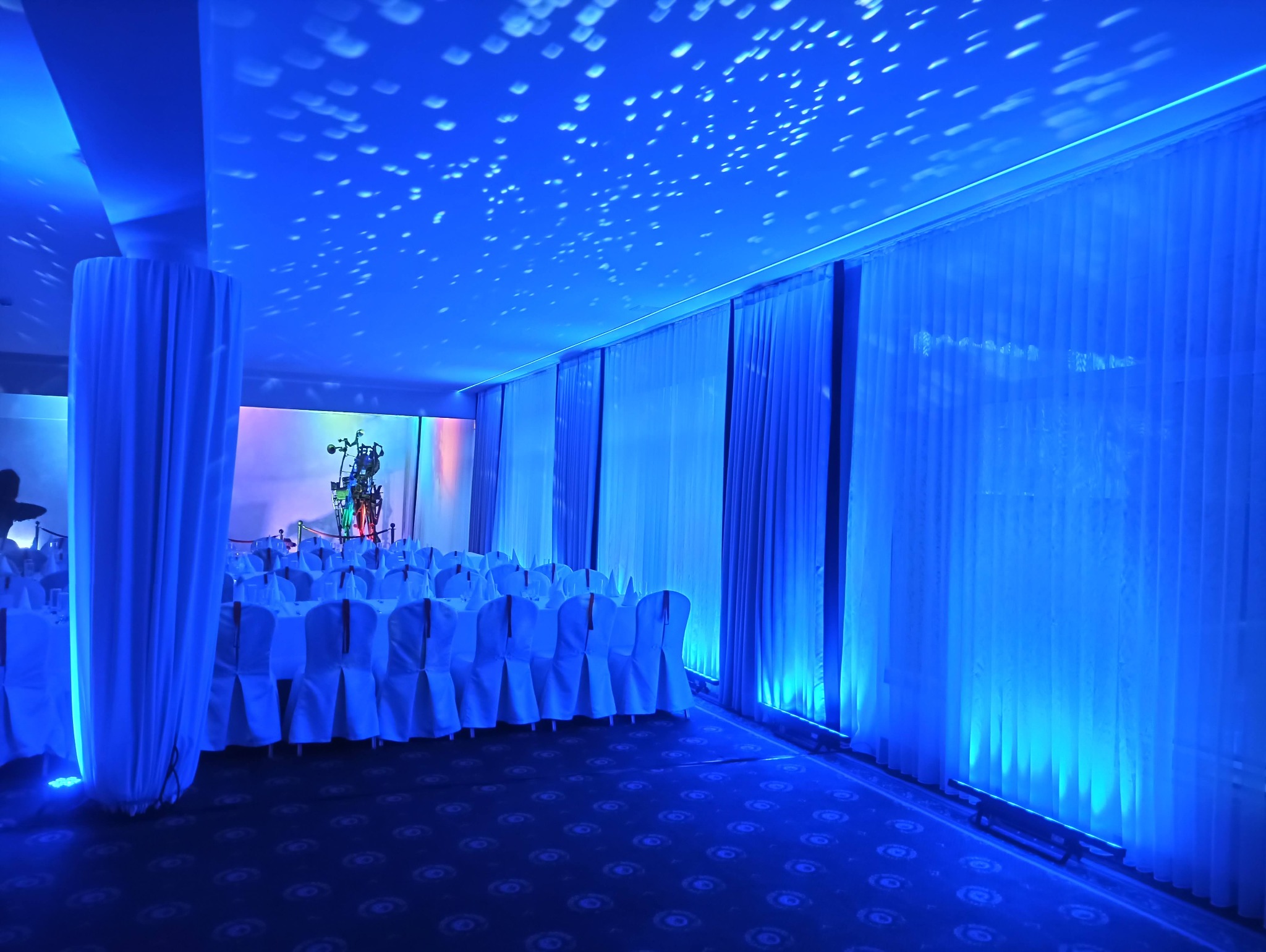 Sala bankietowa przygotowana na uroczystość, oświetlona niebieskim światłem, z dekoracyjnymi zasłonami i stołami z białymi obrusami.