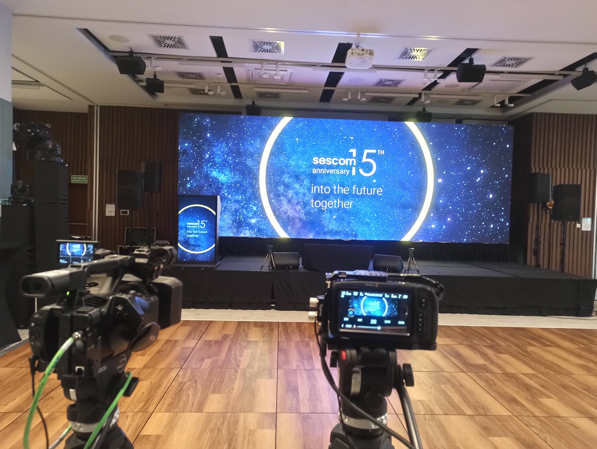 Profesjonalne kamery w studio filmowym ustawione przed sceną z dużym ekranem LED wyświetlającym logo firmy i hasło '15th anniversary into the future together'. Widoczne dodatkowe głośniki...