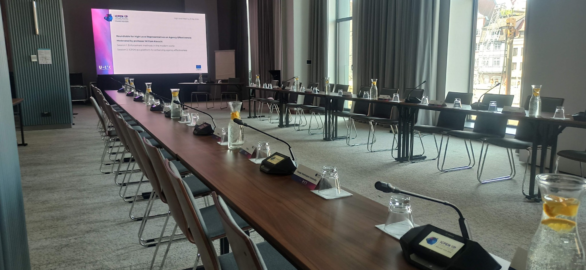 Sala konferencyjna z długim stołem, mikrofonami, szklankami i dzbankami z wodą z cytryną, przygotowana na spotkanie. W tle ekran z prezentacją i widok na budynki za oknem.
