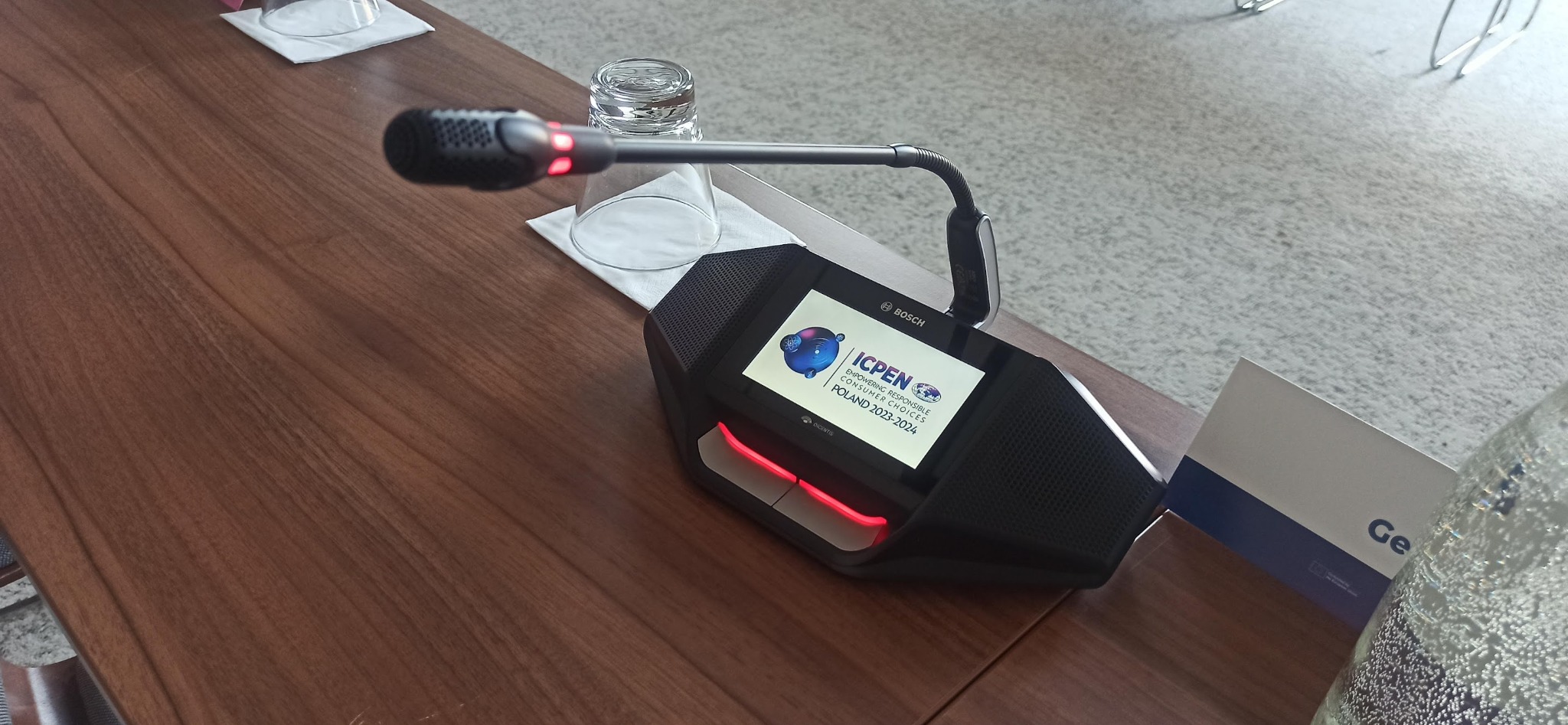 Czarny mikrofon konferencyjny Bosch z ekranem wyświetlającym logo ICPEN na drewnianym stole, obok szklanka z wodą.