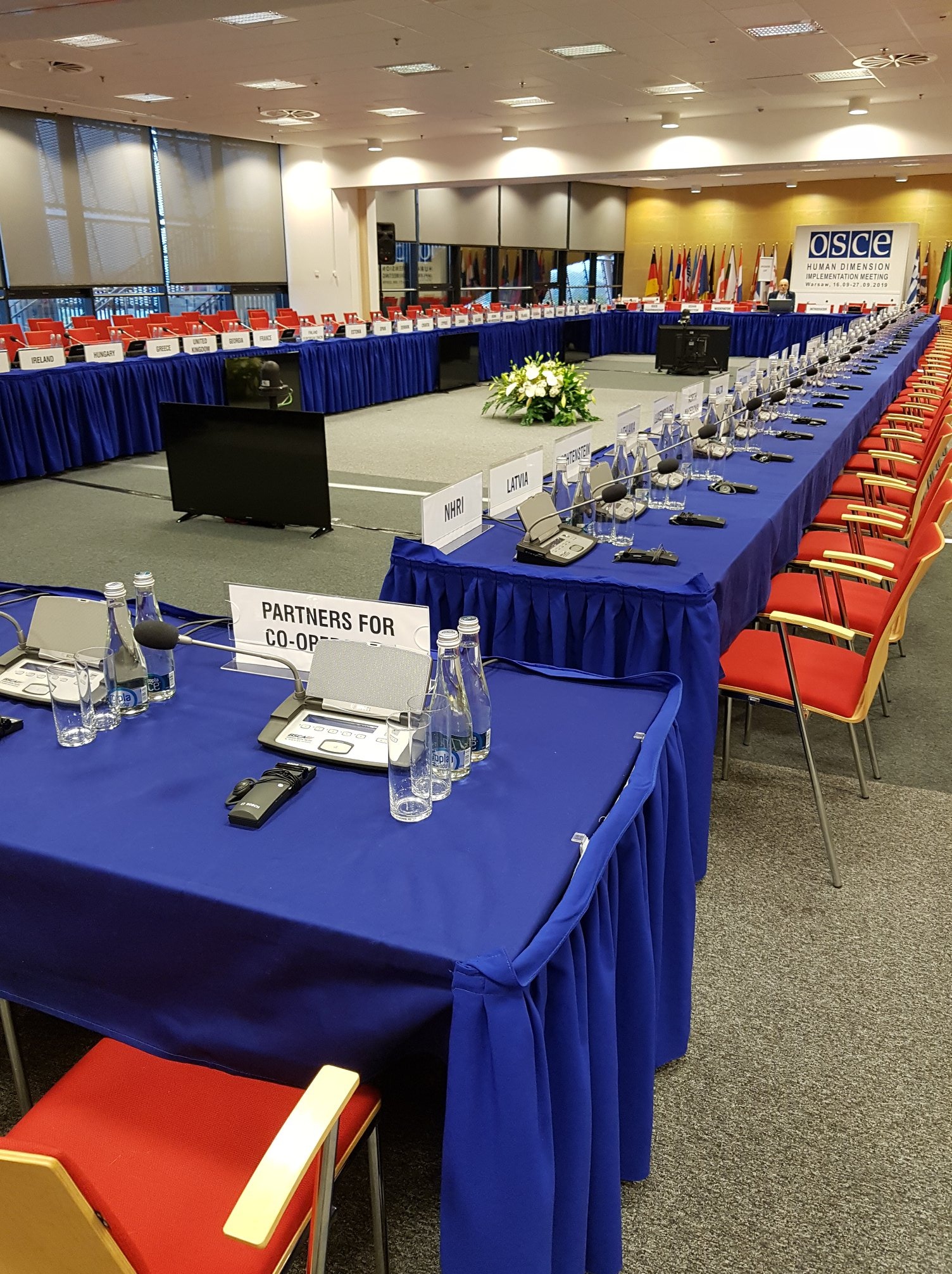 Sala konferencyjna z długim stołem przykrytym niebieskim obrusem, krzesła w kolorze czerwonym, mikrofony, szklanki z wodą i tabliczki z nazwami państw.