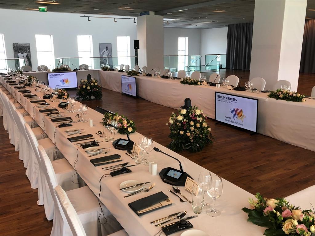 Długi stół konferencyjny z białym obrusem, nakryty dla wielu osób, z mikrofonami, szklankami, sztućcami i kwiatowymi dekoracjami; w tle ekrany wyświetlające napis 'Berlin Process'.