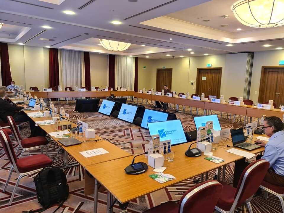 Sala konferencyjna w Gdyni, przygotowana do wydarzenia z monitorami wyświetlającymi logo, ustawionymi na stołach w kształcie podkowy. Widoczni uczestnicy i sprzęt konferencyjny.