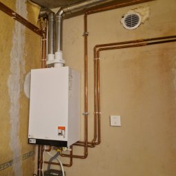 TK-SERWIS Kamil Tymiak - Biały piec gazowy Viessmann z miedzianą instalacją, rurami odprowadzającymi spaliny i wentylacją na ścianie w pomieszczeniu z surowym wykończeniem.