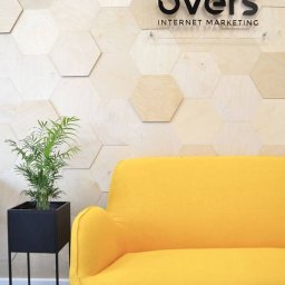 Overs sp. z o.o. - Recepcja firmy Over's Internet Marketing z żółtą sofą, rośliną w czarnej donicy i ścianą z drewnianych heksagonów.