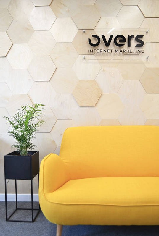 Recepcja firmy Over's Internet Marketing z żółtą sofą, rośliną w czarnej donicy i ścianą z drewnianych heksagonów.