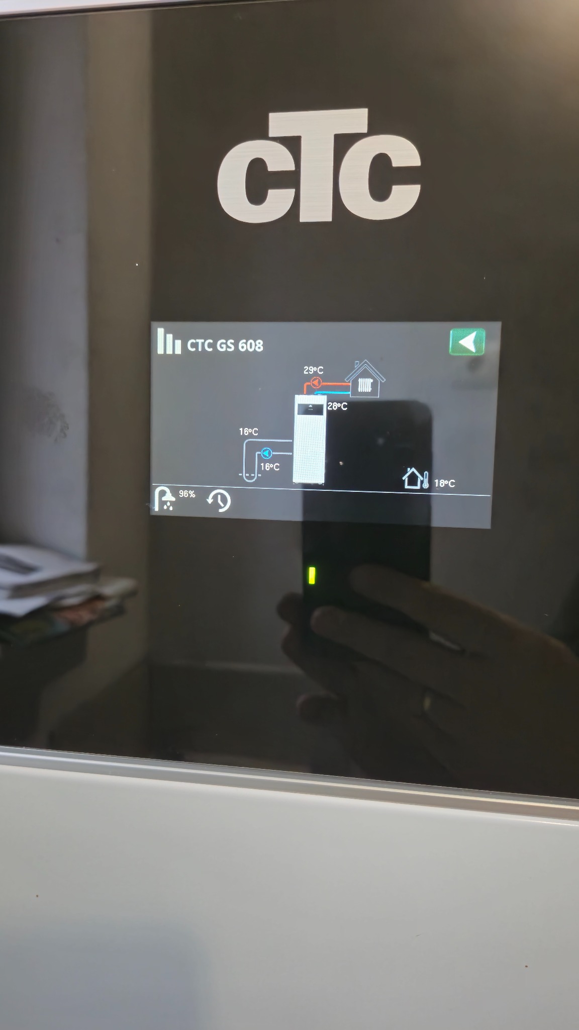 Ekran sterownika pompy ciepła CTC GS 608 z widocznymi temperaturami i schematem instalacji grzewczej, odbicie dłoni na błyszczącej powierzchni.