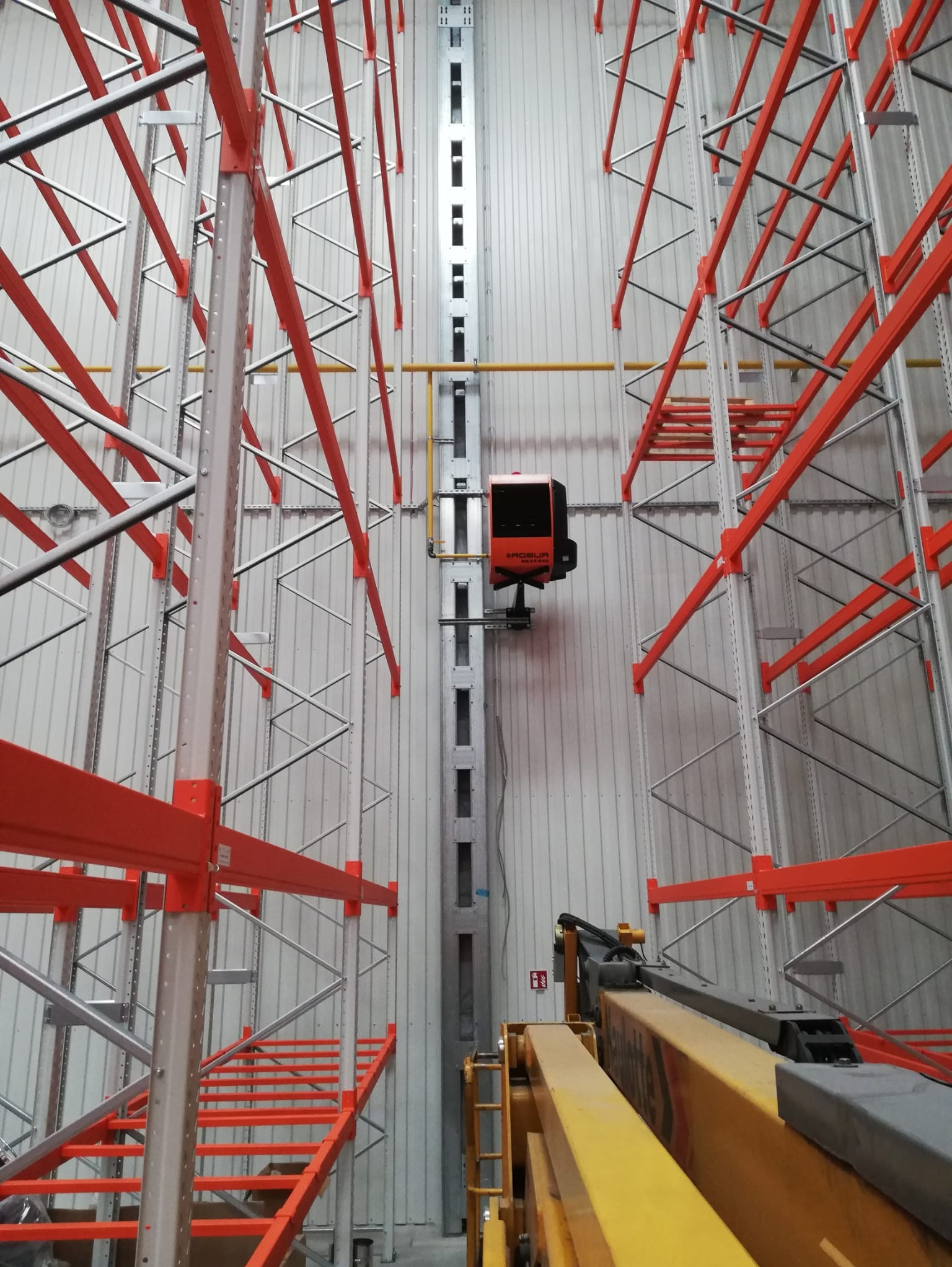 Wysokie regały magazynowe z pomarańczowymi belkami, widok z dołu z wózkiem widłowym z przodu i urządzeniem grzewczym na jednej z półek w tle.