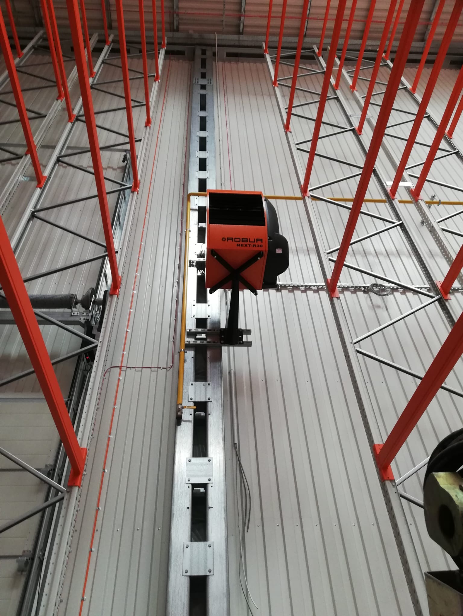 Widok z dołu na zamontowany rekuperator Robur NEXT-R30 na szynie w hali przemysłowej, widoczne pomarańczowe elementy konstrukcyjne i instalacje.