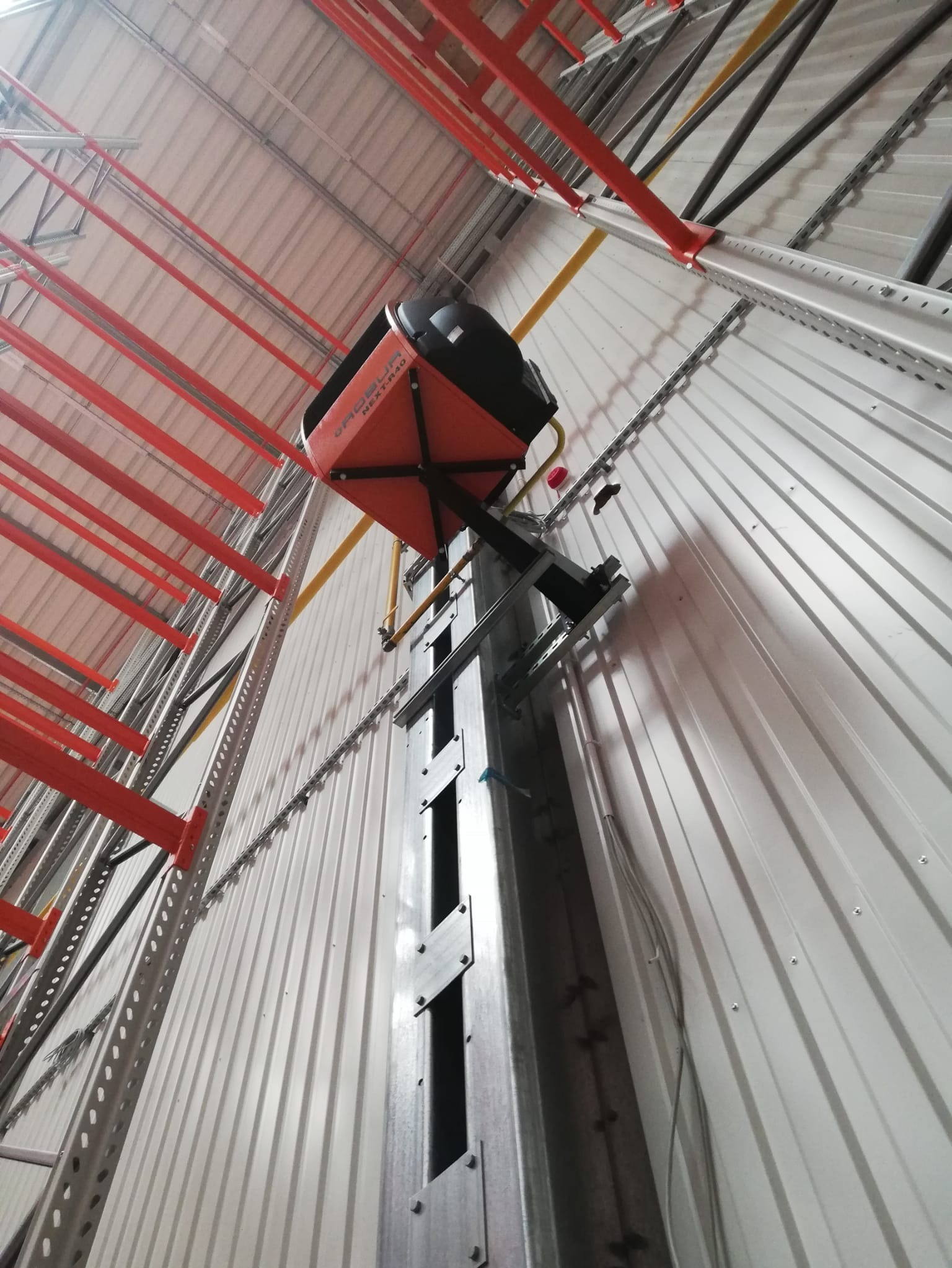 Pomarańczowo-czarna jednostka rekuperacyjna zamontowana na metalowej konstrukcji przy ścianie z blachy trapezowej w hali przemysłowej, widok z dołu.