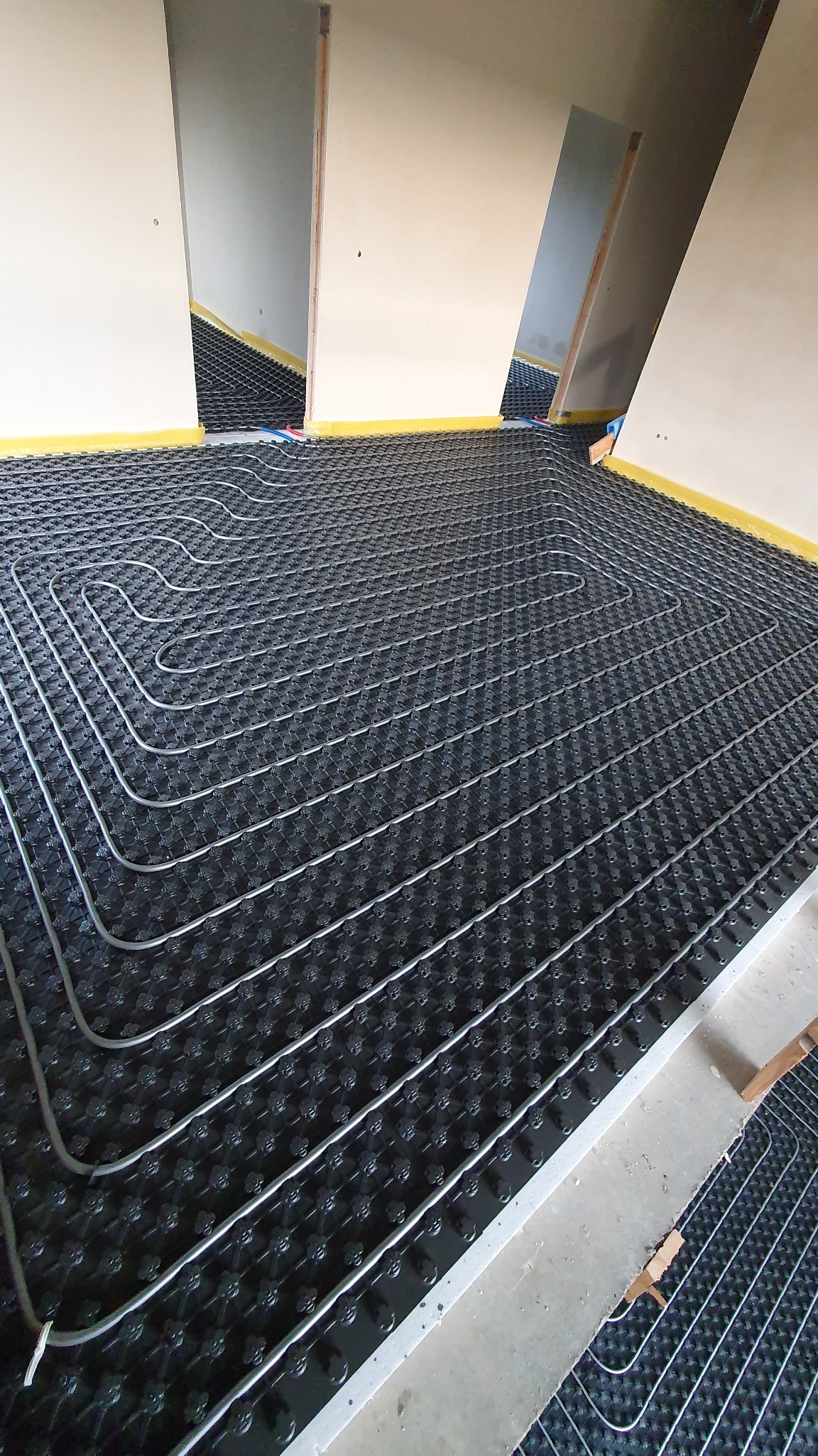 Instalacja ogrzewania podłogowego w trakcie montażu, widoczne rury grzewcze ułożone na czarnej macie izolacyjnej z wypustkami, żółta taśma wzdłuż ścian.