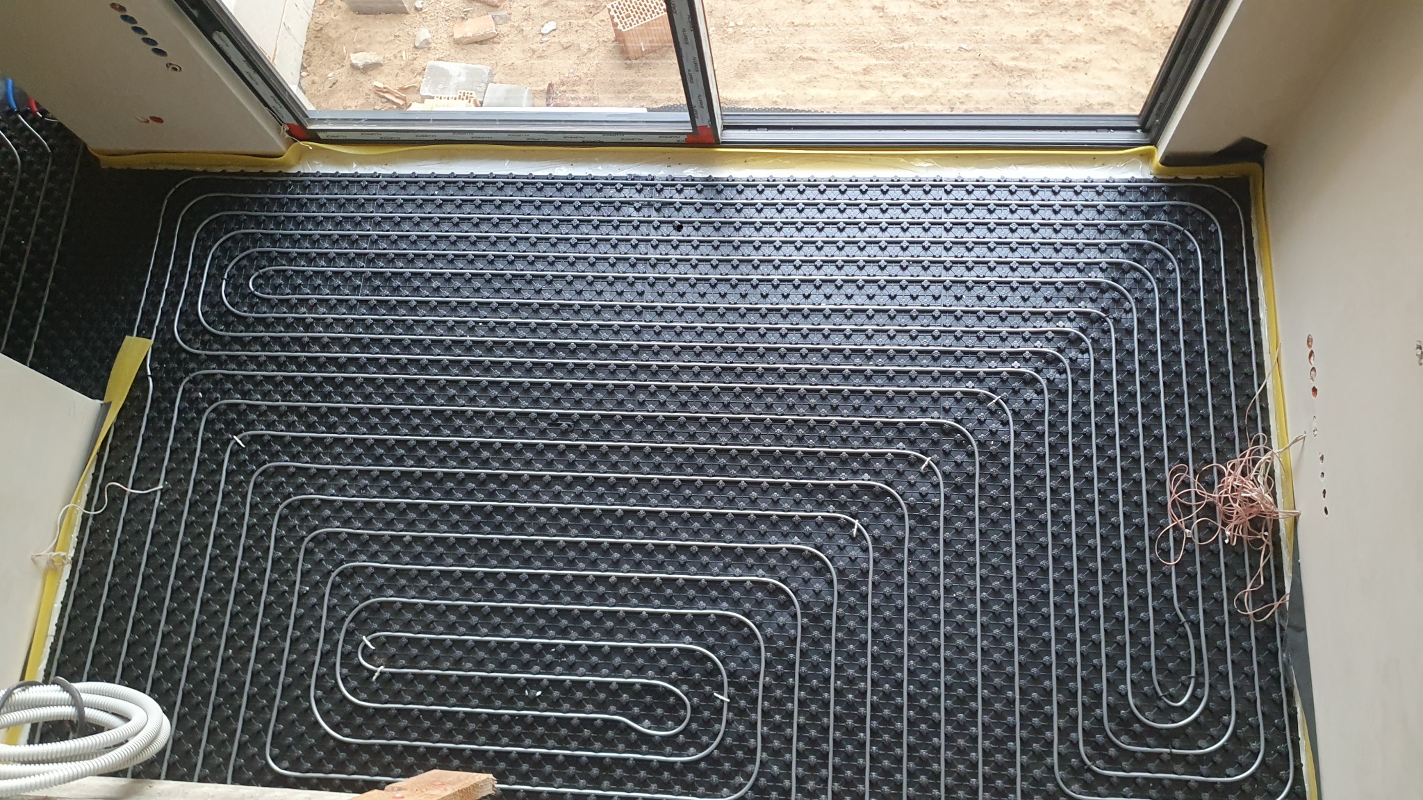 Widok z góry na rozłożone rury ogrzewania podłogowego na czarnej macie izolacyjnej w trakcie instalacji, widoczne kable elektryczne przy ścianie i fragment okna balkonowego.