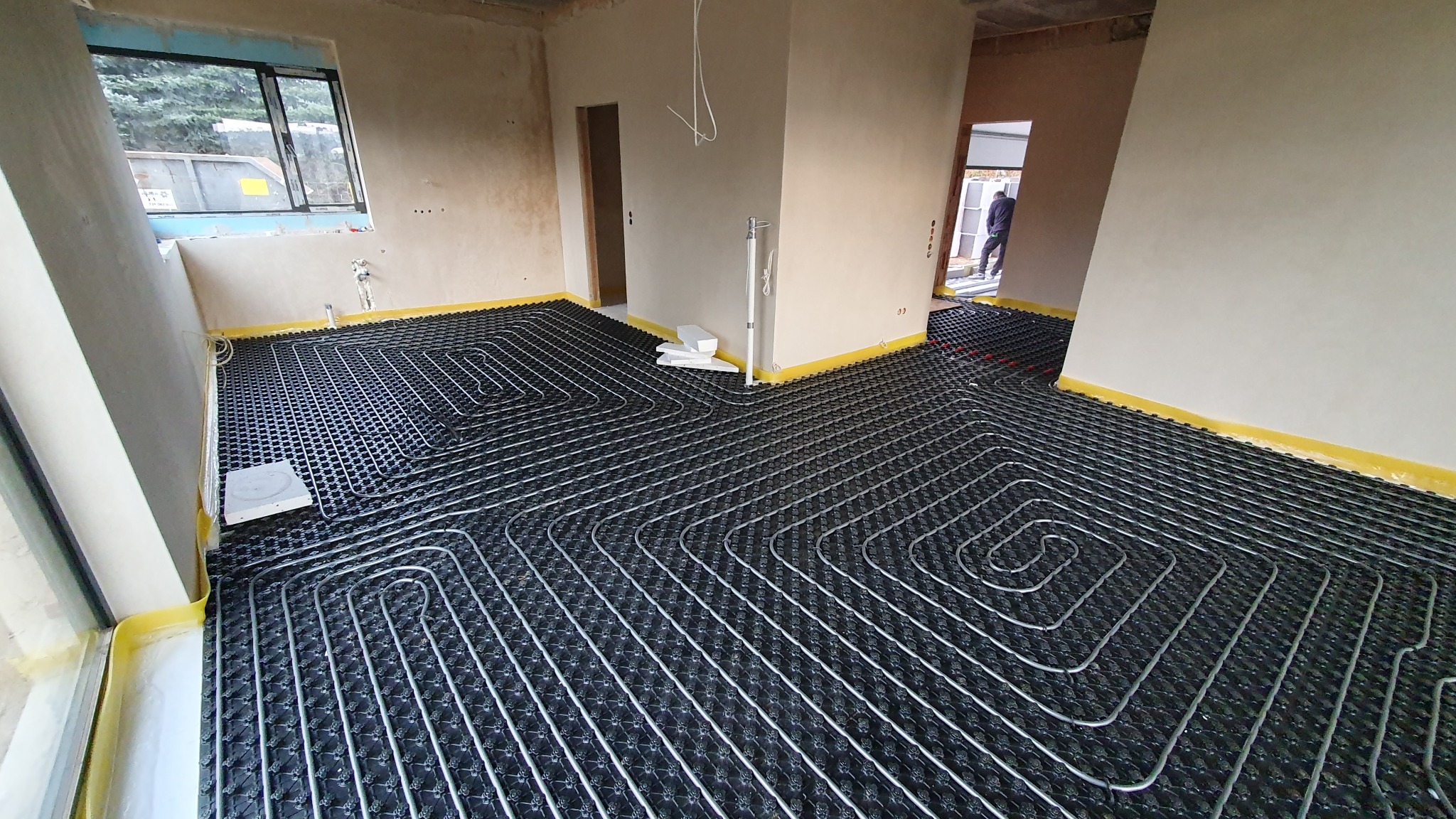 Rozłożona instalacja ogrzewania podłogowego w surowym wnętrzu, widoczne rury grzewcze ułożone na czarnej macie z żółtą taśmą przy ścianach, okno z widokiem na zewnątrz i częściowo widoczna osoba...