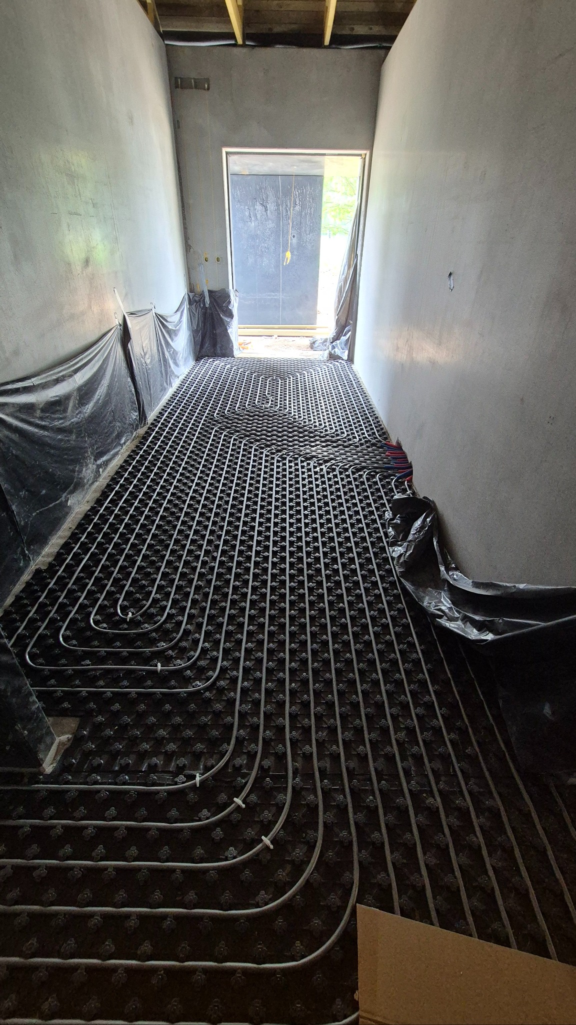Rozłożone rury ogrzewania podłogowego na czarnej macie izolacyjnej w surowym wnętrzu budynku, widok w perspektywie w kierunku drzwi na zewnątrz.