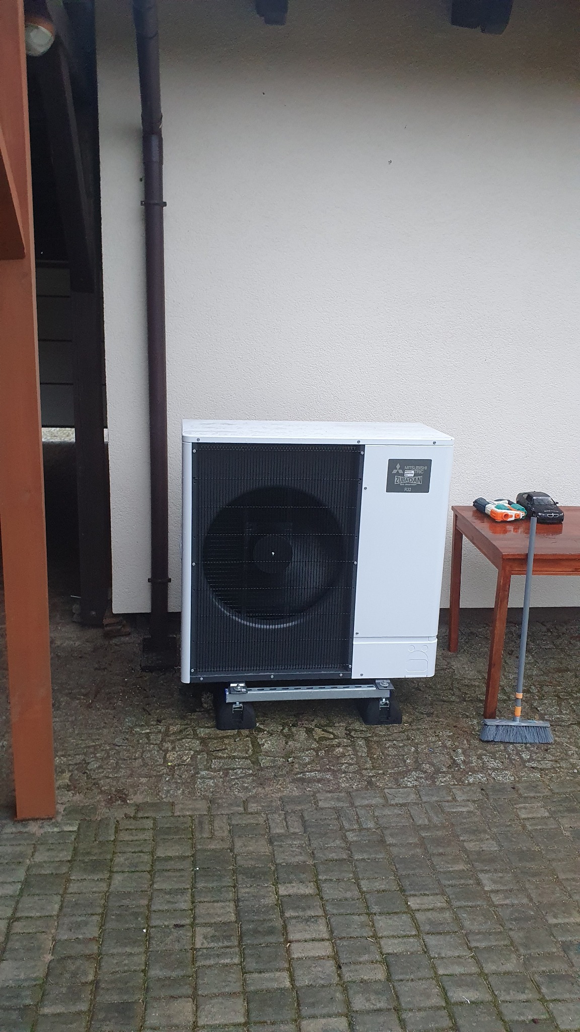 Biała pompa ciepła Mitsubishi Zubadan R32, zamontowana na metalowej podstawie na kostce brukowej, obok drewniany stolik z zabawkami i miotłą, ściana budynku z rynną w tle.