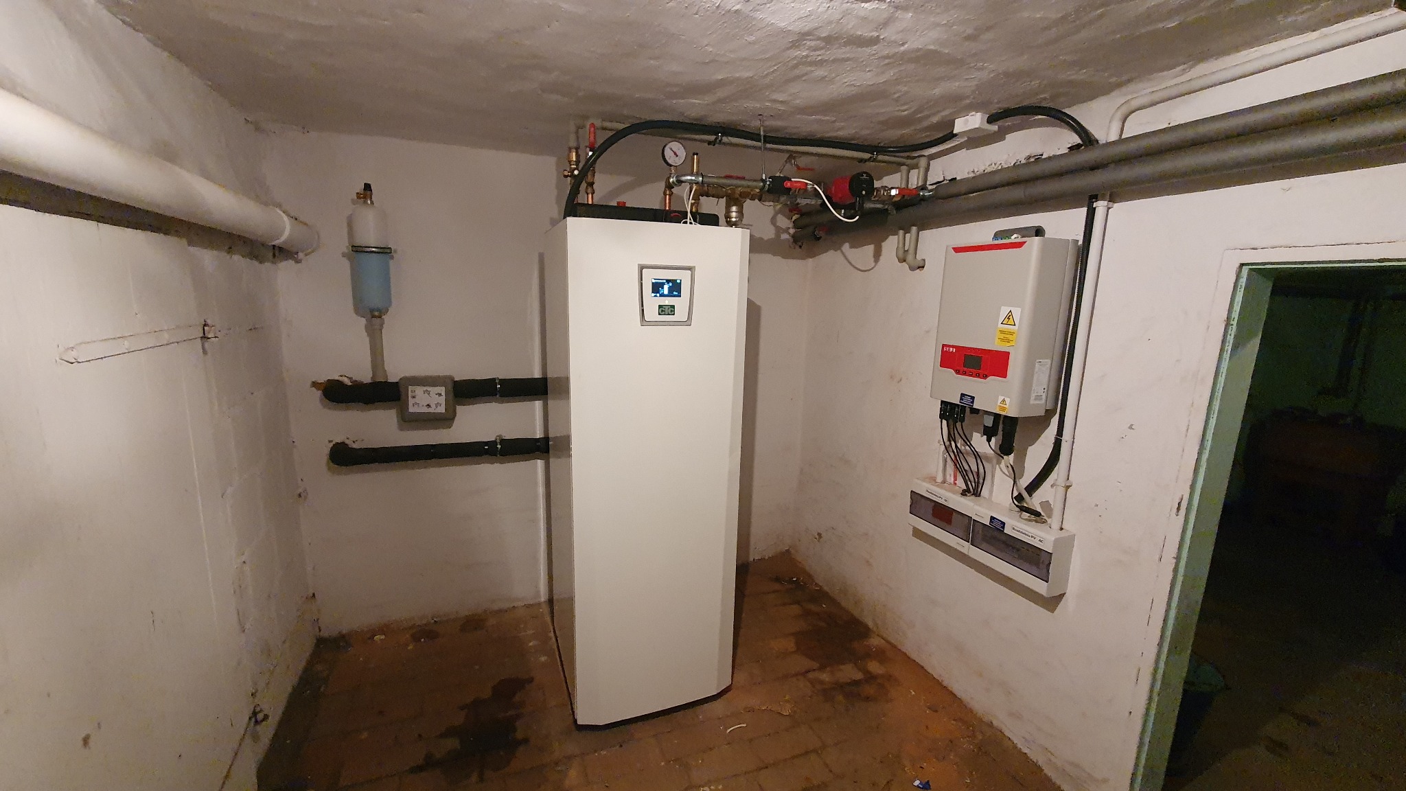 Biała pompa ciepła z wyświetlaczem w piwnicy, widoczne rury, zawory, manometr oraz falownik na ścianie obok drzwi.