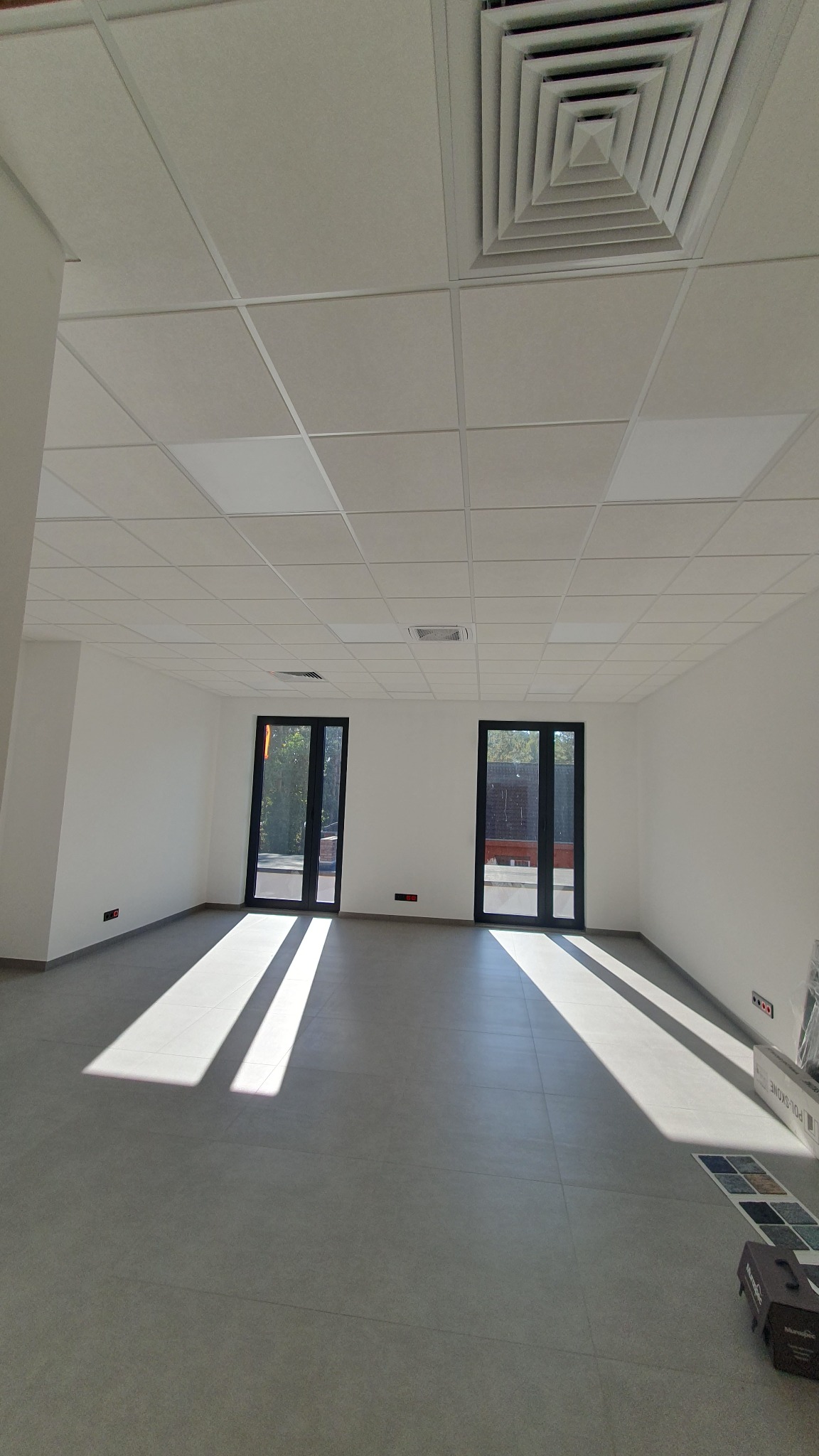 Puste pomieszczenie z szarą podłogą, białymi ścianami i dwoma czarnymi oknami, wpadające promienie słoneczne, sufit kasetonowy z kratką wentylacyjną, próbki płytek podłogowych przy ścianie.