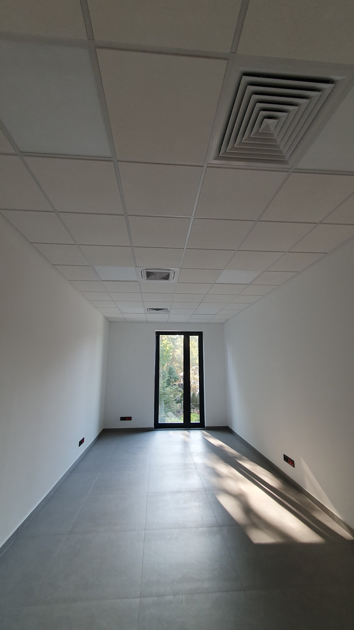 Wnętrze pustego pomieszczenia z szarą podłogą z płytek, białymi ścianami i sufitem podwieszanym z kratką oraz kratką wentylacyjną, widok na drzwi balkonowe z widokiem na zieleń na zewnątrz...
