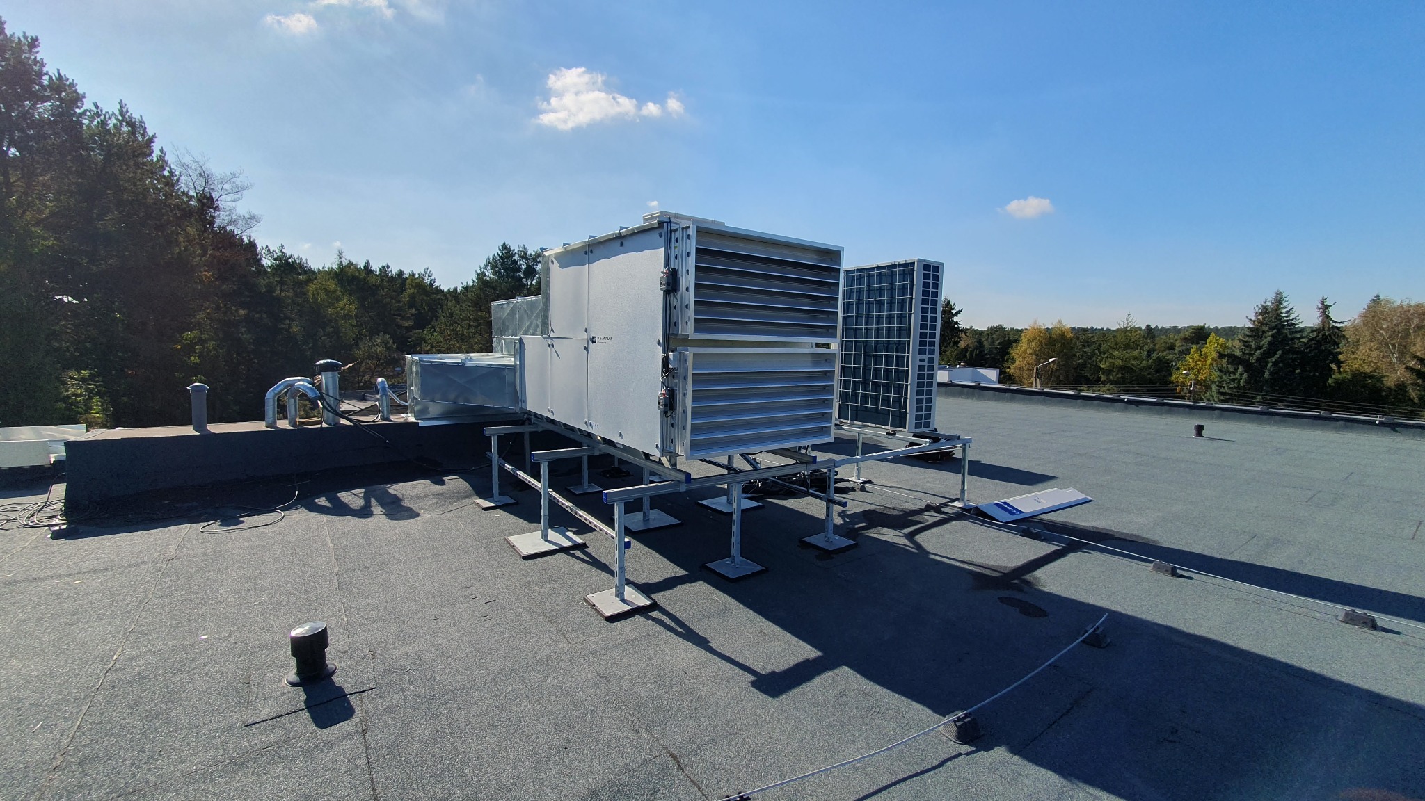 Instalacja rekuperacji na dachu budynku, widoczne jednostki centralne z wentylatorami i systemem kanałów wentylacyjnych, zamontowane na metalowej konstrukcji wsporczej, w tle las i błękitne niebo.