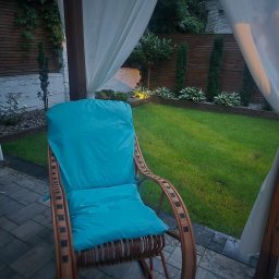 Syla Decor - Wiklinowy fotel bujany z turkusową poduszką w ogrodzie w Kiekrz. Meble ogrodowe z nowym obiciem, widoczne białe zasłony i trawnik.