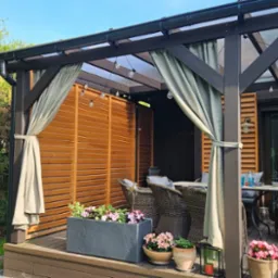 Zadaszony taras z drewnianą konstrukcją, zasłonami w kolorze ecru i kwiatami w donicach. Elementy dekoracyjne i oświetlenie tworzą przytulny klimat.