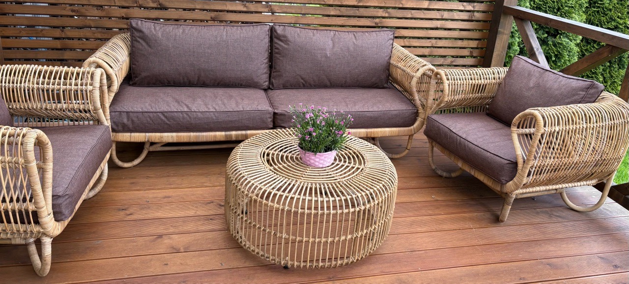 Komplet mebli ogrodowych z rattanu: sofa, dwa fotele i stolik kawowy z kwiatami w doniczce. Brązowe poduszki na siedziskach i oparciach. Drewniana podłoga tarasu.