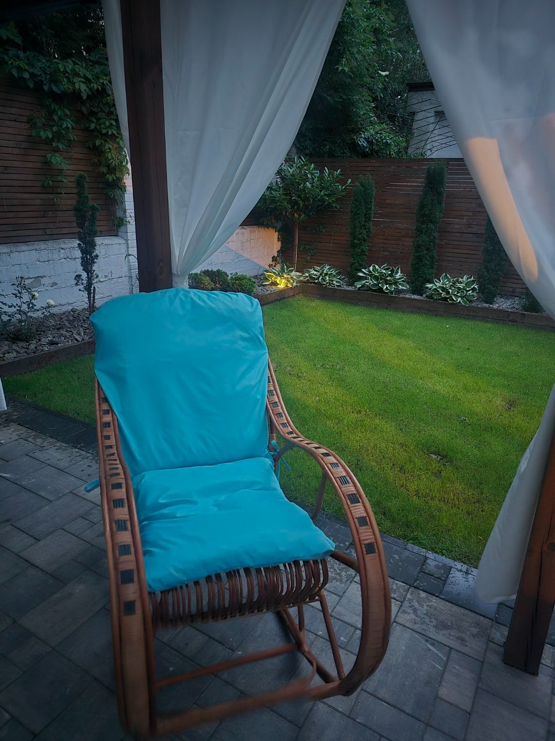 Wiklinowy fotel bujany z turkusową poduszką w ogrodzie w Kiekrz. Meble ogrodowe z nowym obiciem, widoczne białe zasłony i trawnik.