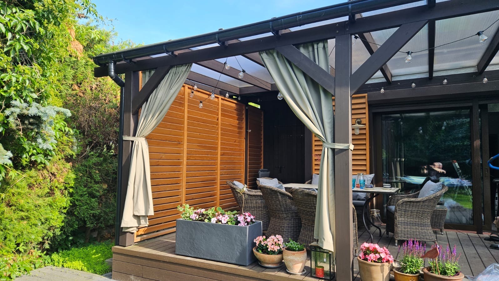 Zadaszony taras z drewnianą konstrukcją, zasłonami w kolorze ecru i kwiatami w donicach. Elementy dekoracyjne i oświetlenie tworzą przytulny klimat.