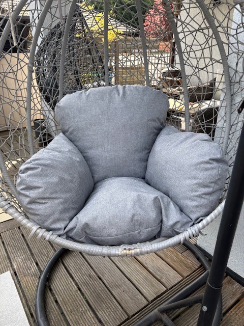 Szary, wiszący fotel kokon z poduszkami na drewnianym tarasie. Widoczne detale plecionki i metalowej konstrukcji. Częściowo zacieniony, letni dzień.