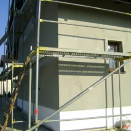 Ściana domu w trakcie ocieplania, widoczne rusztowanie z drewnianą drabiną opartą o nie, fragment okna i wiadro z zaprawą przy dolnej krawędzi ściany.