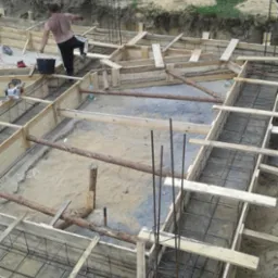 Budowa fundamentów domu: drewniane szalunki zbrojone stalą, widoczny pracownik z wiadrem w trakcie pracy, teren budowy z ogrodzeniem w tle.