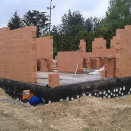 Wznoszenie ścian parteru z czerwonej ceramiki poryzowanej na fundamencie z hydroizolacją, widoczny drenaż fundamentów oraz niebieska rura kanalizacyjna z pomarańczowym zabezpieczeniem.