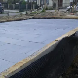 Przygotowanie fundamentów pod budynek: izolacja pozioma z szarego styropianu ułożona na gruncie, zabezpieczona folią kubełkową przy ścianach fundamentowych, widoczny szalunek z drewna, w tle dom...