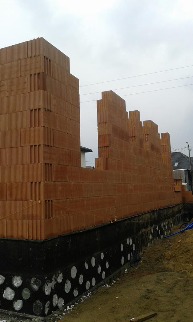 Wznoszenie ścian domu z bloczków ceramicznych Porotherm na fundamencie z izolacją przeciwwilgociową, widoczne zbrojenie i fragmenty sąsiednich budynków.
