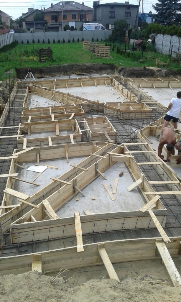 Zbrojenie i szalunek fundamentów pod dom jednorodzinny z widocznym geodetą i paletami w tle.