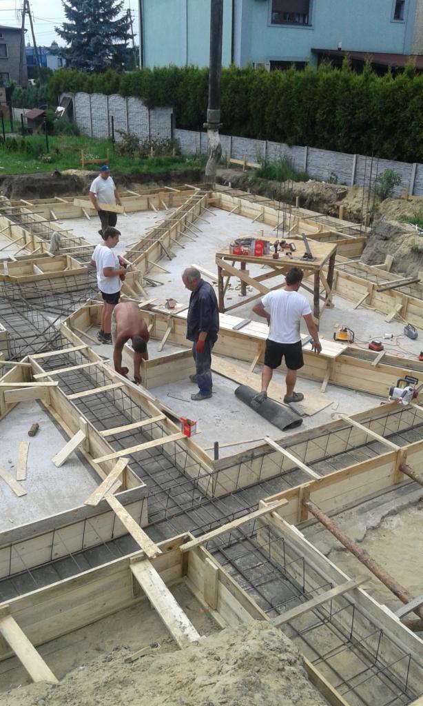 Prace fundamentowe: drewniane szalunki zbrojone stalową siatką na placu budowy, widoczni pracownicy i pompa do betonu w tle.