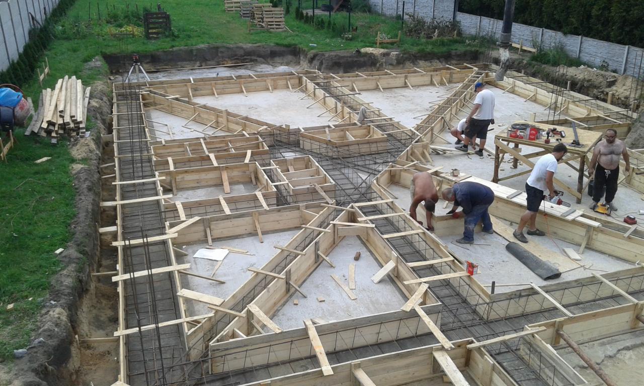 Wykopane fundamenty z drewnianym szalunkiem i zbrojeniem stalowym, ekipa budowlana przy pracy, widok z góry na plac budowy z narzędziami i materiałami budowlanymi.