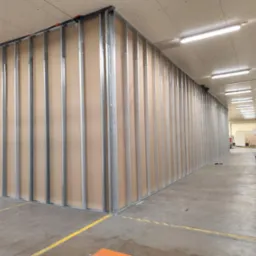 Konstrukcja ścian działowych z płyt kartonowo-gipsowych na metalowym stelażu w dużej hali. Widoczne profile i płyty. Żółte linie na posadzce.