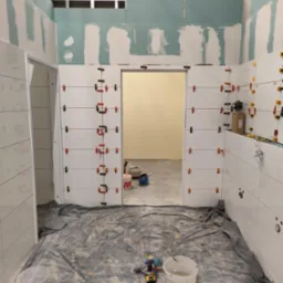 Białe płytki układane w łazience, z użyciem klipsów poziomujących. Widoczne narzędzia i folia ochronna na podłodze. Ściany częściowo wykończone płytami gipsowo-kartonowymi.