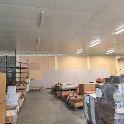 PERFECT-BUD - Wnętrze hali w trakcie prac wykończeniowych: ściany z płyt OSB, pracownik w odzieży roboczej, materiały budowlane na paletach. Widoczne oświetlenie sufitowe.