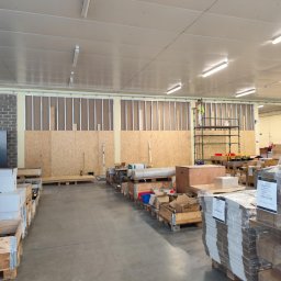 PERFECT-BUD - Duża przestrzeń magazynowa w trakcie prac wykończeniowych: montaż płyt OSB i konstrukcji stalowej. Pracownik na rusztowaniu. Materiały budowlane w paletach.