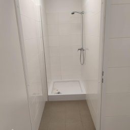 PERFECT-BUD - Wykończona kabina prysznicowa z białymi płytkami i brodzikiem, widoczna przez otwarte drzwi. Chromowana bateria i słuchawka prysznicowa. Podłoga wyłożona brązowymi płytkami.