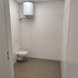 PERFECT-BUD - Jasna łazienka z białymi płytkami, toaletą i bojlerem. Brązowa podłoga z płytek. Widok przez otwarte drzwi. Minimalistyczny design.