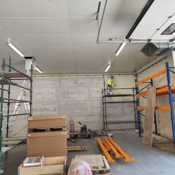 PERFECT-BUD - Prace wykończeniowe w hali: pracownik na rusztowaniu szpachluje ścianę. Widoczne materiały budowlane, regał magazynowy i znak informacyjny.