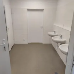 Wyremontowana łazienka publiczna z trzema umywalkami, jasnymi płytkami na ścianach i beżową podłogą. Widoczne drzwi i oświetlenie sufitowe.